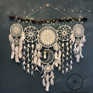 May include: A white dreamcatcher wall hanging with a crescent moon and star design. The dreamcatcher is made of white feathers and beads. The dreamcatcher is hanging from a branch with white lights.