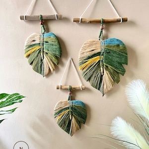 Könnte beinhalten: Drei Makramee-Wandbehänge in Form von Monstera-Blättern. Die Blätter sind aus Naturfasern in Grün-, Beige- und Blautönen gefertigt. Jeder Behang ist an einem Holzstab und einer weißen Schnur aufgehängt.