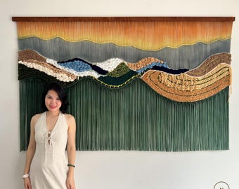 May include: A large, woven wall hanging depicting a landscape scene. The artwork features a gradient of colors, from orange and yellow at the top to green at the bottom, with textured, layered elements in between. The piece is mounted on a wooden frame.