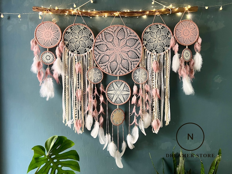 May include: A set of seven pink and white dreamcatchers with feathers hanging from a wooden branch. The dreamcatchers are arranged in a row and are all different sizes. The dreamcatchers are made of macrame and have a delicate, bohemian style.