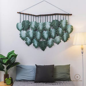 May include: A green macrame wall hanging with a wooden dowel at the top. The wall hanging is made of a series of knotted leaves, creating a tropical theme.