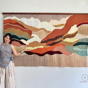 May include: A large rectangular wall hanging featuring a landscape design made of textured yarn in shades of burnt orange, cream, dark green, mustard yellow, and tan. The piece is mounted on a dark wooden rod and includes long fringe at the bottom.