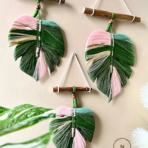 May include: Three macrame leaf wall hangings with pink, green and beige yarn. The leaves are hanging from wooden dowels with string. The text "DREAMER STORE" is visible in the bottom right corner of the image.