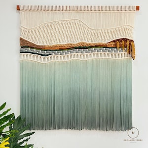 以下が含まれることがあります： 上部に木の棒が付いた、長方形の大きなマクラメの壁掛け。クリーム色、茶色、緑色、灰色のテクスチャの層があり、下部には緑色と灰色のグラデーションのフリンジがあります。Dreamer Storeのロゴが右下にあります。