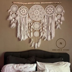 May include: A white dream catcher wall hanging with multiple layers of crocheted circles and feathers. The dream catcher is hanging on a branch above a black headboard with white bedding.