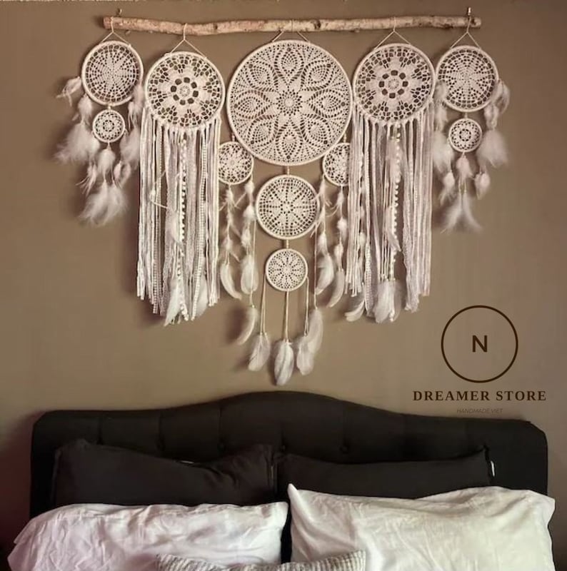 May include: A white dream catcher wall hanging with multiple layers of crocheted circles and feathers. The dream catcher is hanging on a branch above a black headboard with white bedding.