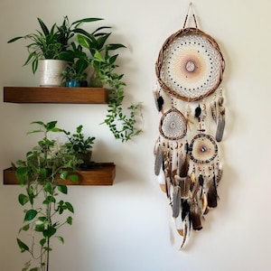 May include: A large, handcrafted dreamcatcher with a woven brown willow frame and a central web design. It features multiple smaller dreamcatchers and is adorned with feathers in shades of brown, black, and white. The dreamcatcher hangs on a white wall.