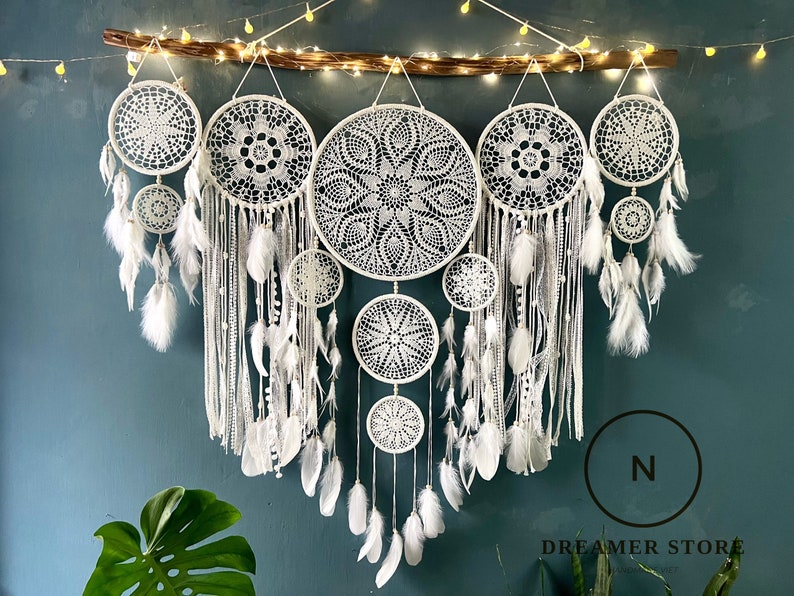 May include: A set of seven white dreamcatchers of varying sizes, hanging from a wooden branch with white feathers and beads. The dreamcatchers are arranged in a cascading pattern, with the largest dreamcatcher at the top and the smallest at the bottom. The dreamcatchers are all made with intricate crocheted designs.