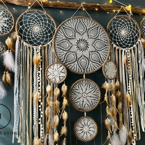 May include: A set of seven white and brown dreamcatchers with feathers and beads hanging from a wooden branch. The dreamcatchers are arranged in a row, with the largest one in the centre. The dreamcatchers are made of crocheted lace and have a rustic, bohemian style.