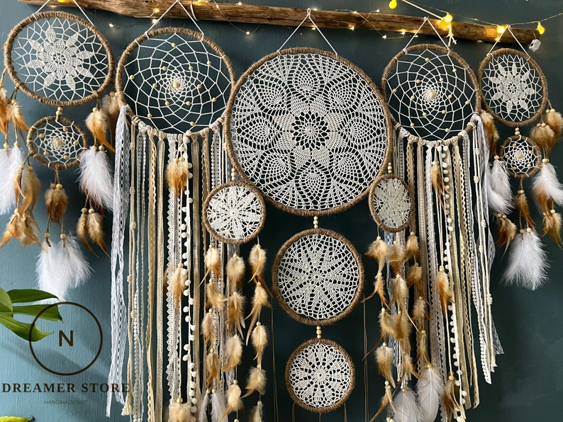 May include: A set of seven white and brown dreamcatchers with feathers and beads hanging from a wooden branch. The dreamcatchers are arranged in a row, with the largest one in the centre. The dreamcatchers are made of crocheted lace and have a rustic, bohemian style.