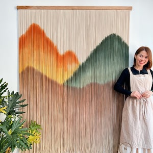 Puede incluir: Un gran tapiz de pared rectangular con un diseño de montaña. La obra de arte presenta un marco de madera y hebras verticales en tonos beige, naranja y verde. La parte inferior de la obra es de color marrón claro. El texto "DREAMER STORE" es visible.