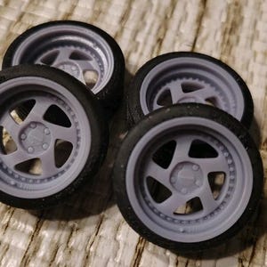 May include: Four gray model car wheels with black tires are arranged on a light brown surface. The wheels have a five-spoke design and a detailed rim. The tires have a realistic tread pattern.