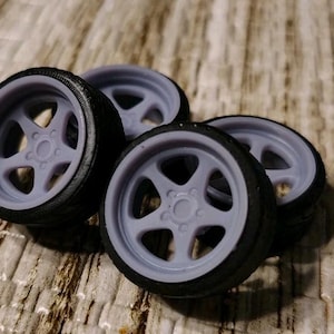 May include: Four miniature car wheels with gray rims and black tires are arranged on a textured surface. The wheels have a five-spoke design and are likely for a model car or similar project.