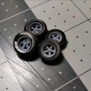 May include: A set of three miniature model car wheels featuring black rubber tires and light gray five-spoke rims. The wheels are arranged in a cluster, showcasing the detailed tread pattern on the tires and the central hub design of the rims.