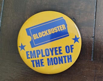 May include: A round, yellow pin with the Blockbuster logo in blue, along with the text "EMPLOYEE OF THE MONTH" in blue. Two blue stars flank the text. The pin is a retro-themed item.