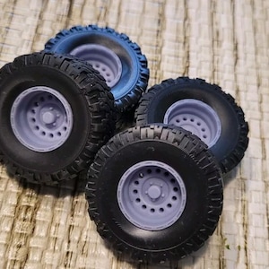 May include: Four black off-road tires with gray rims are arranged on a textured surface. The tires have a deep tread pattern, and the rims feature multiple circular holes. The tires are likely for a model vehicle.