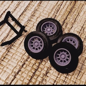 May include: Four black toy car tires with light purple rims are arranged on a textured surface. A black, angular frame is positioned to the left of the tires. The tires have a diameter of approximately 1.5 inches.