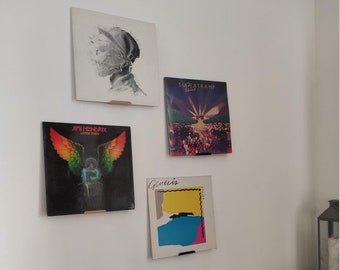 May include: Four vinyl records displayed on a white wall using black and brown wall mounts. The records are by Jimi Hendrix, Supertramp, Genesis, and an unknown artist. The records are displayed in a staggered pattern.
