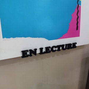 Può includere: Una cornice bianca con una stampa astratta colorata e le parole "EN LECTURE" in lettere nere attaccate alla parte inferiore.