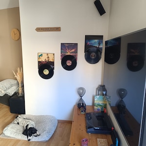 May include: A living room scene featuring vinyl records displayed on a white wall. A wooden sign reads "LA REUNION 9342 KM". A dog rests on a fluffy bed. A wooden clock and a vase with dried plants are also visible.