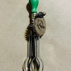May include: Vintage hand-crank egg beater with a green wooden handle. The beater has a metal gear mechanism and stainless steel whisk blades. The beater is a kitchen utensil.