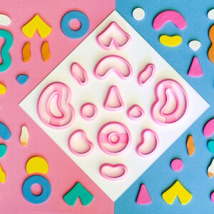 以下が含まれることがあります： ドーナツ、ハート、三日月など、さまざまな形をしたピンクのクッキーカッターのセット。カッターは白い背景に配置され、カラフルな形に囲まれています。