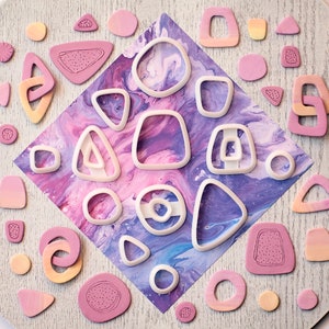 May include: A collection of pink and white plastic cookie cutters in various shapes, including circles, triangles, and squares, arranged on a light wood surface.
