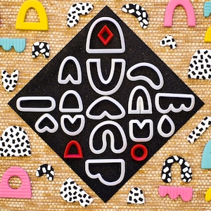 以下が含まれることがあります： 黒地にカラフルな幾何学模様が施された背景に、アーチ、ハート、渦巻きなど、さまざまな形をした白いプラスチック製のクッキー型18個のセットです。