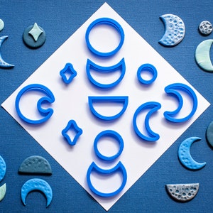 Peut inclure: Un ensemble de 10 emporte-pièces en plastique bleu en forme de lunes, d'étoiles et de cercles. Les emporte-pièces sont disposés sur un fond blanc avec des formes en pâte polymère bleue autour des bords.
