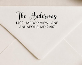 May include: A white envelope featuring black printed text that reads The Andersons, 1492 Harbor View Lane, Annapolis, MD 21401. The envelope is positioned near a wooden rubber stamp with a black handle, showcasing a clean and professional design.