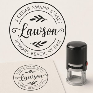 May include: A black self-inking round stamp featuring the name Lawson in a script font, surrounded by the address 5 Cedar Swamp Street, Howard Beach, NY 11414, with two small leaf illustrations above and below the name, shown with a sample imprint.