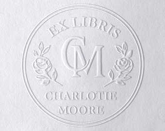 May include: A white embossed design featuring the words "EX LIBRIS" at the top, the initials "CM" in the center, and the name "CHARLOTTE MOORE" at the bottom. The design includes floral elements and is circular.