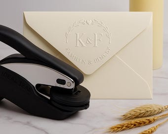 May include: A cream-colored envelope with embossed initials "K & F" inside a wreath, with the names "Kamryn & Finley" below. A black and silver Ideal Seal embosser is in the foreground. Wheat stalks and a candle are in the background.
