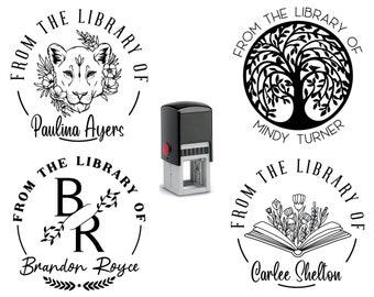May include: A black and gray self-inking stamp with four different circular designs. Each design includes the text "FROM THE LIBRARY OF" and a name. The designs feature a lion, a tree, initials, and an open book with flowers.