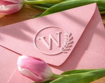 May include: A pink envelope with a debossed letter "W" inside a circle, accompanied by a sprig of leaves. The envelope is set against a wooden surface, with pink and white tulips and green leaves.
