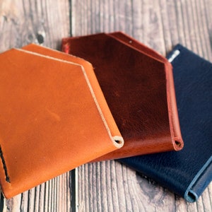 Può includere: Sono mostrati tre porta carte in pelle nei colori arancione, marrone e blu navy. Ogni portafoglio ha un design minimalista con un bordo cucito. I porta carte sono disposti su una superficie di legno.