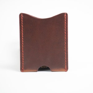 May include: A brown leather card holder with red stitching. The card holder has a curved top and a cutout at the bottom. The leather has a slightly textured appearance.
