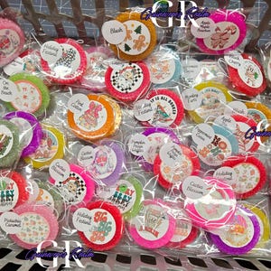 May include: A basket filled with various round, colorful, and scented air fresheners. Each air freshener is individually packaged and labeled with names like "Pistachio Caramel," "Holiday Festival," and "Cactus Blossom." The labels feature festive graphics.