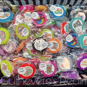 May include: A variety of colorful round car air fresheners with different designs and scents. The scents include "Pink Sugar", "Peach Bellini", "Watermelon Lemonade", "Cactus Blossom", "Creme Brulee", "Let the Good Times Roll", "Vanilla Orange", "Pina Colada", "Frosted Cupcake", "Volcano", "True Loops", "One Love", "Good Vibes", "Drop King", "You've Outgrown It", "Loon Stop", "Shroom Queens", and "Pink Sugar".