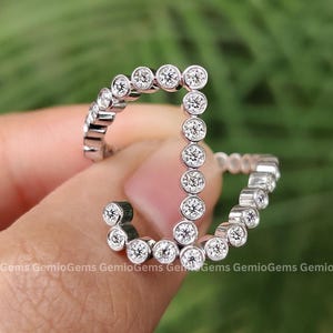 May include: A silver ring with a 'J' shape, encrusted with small, round, clear gemstones. The ring is held between fingers against a blurred green background, showcasing the ring's design and sparkle.