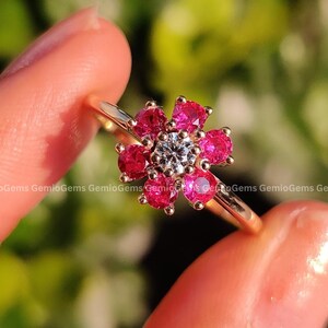 May include: A gold ring featuring a flower design. The flower has a central white gemstone surrounded by vibrant red gemstones. The ring is held against a blurred green background.