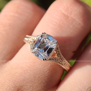 Puede incluir: Un anillo con una gran piedra central transparente de corte cojín montada en una banda de oro rosa. La banda muestra un intrincado trabajo de filigrana grabado en los hombros y un detalle de borde milgrain alrededor del engaste central.
