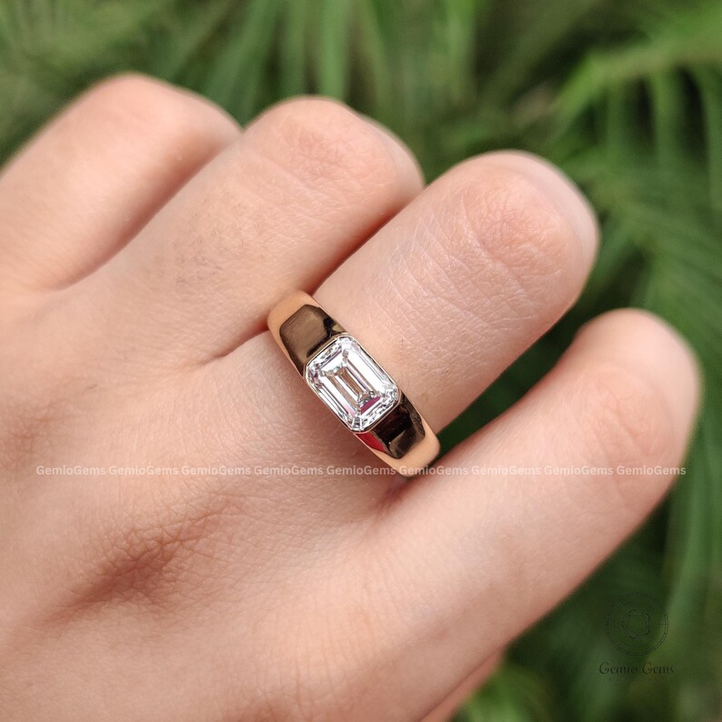 May include: A gold ring featuring a single emerald-cut diamond set in a bezel setting. The band is smooth and polished, showcasing a reflective metallic surface. The center stone is clear and rectangular, positioned horizontally on the finger.