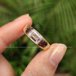 May include: A gold ring featuring a single emerald-cut clear gemstone set in a smooth bezel setting. The band is polished gold with a solid, rounded profile, showcasing a clean and reflective surface that highlights the central rectangular stone.