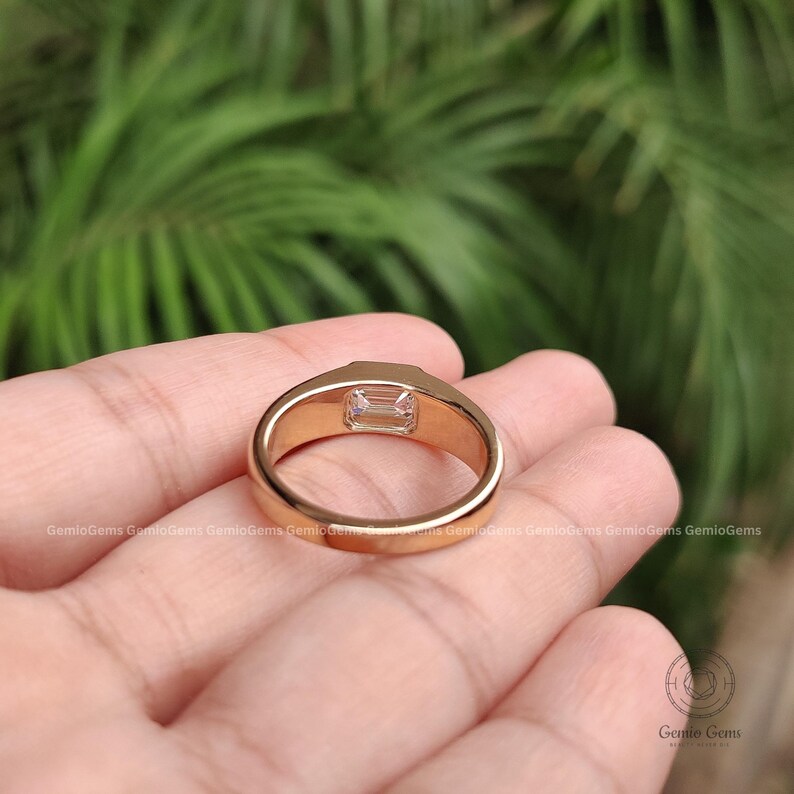 May include: A gold ring featuring a single emerald-cut clear gemstone set in a smooth, polished band. The ring has a solid, thick metal construction with a reflective surface, showcasing the rectangular stone from a side-profile perspective.