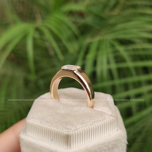 May include: A gold ring featuring a single emerald-cut clear gemstone set in a smooth, polished band. The ring is positioned upright on a white, textured, hexagonal display cushion, showcasing the reflective metal surface and the faceted stone.