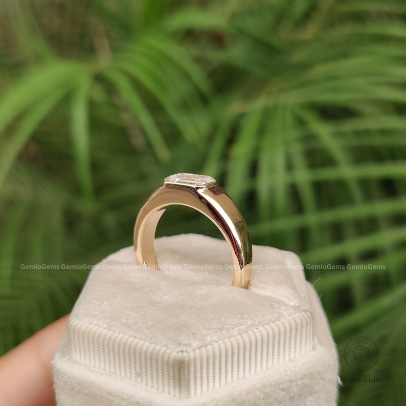 May include: A gold ring featuring a single emerald-cut clear gemstone set in a smooth, polished band. The ring is positioned upright on a white, textured, hexagonal display cushion, showcasing the reflective metal surface and the faceted stone.