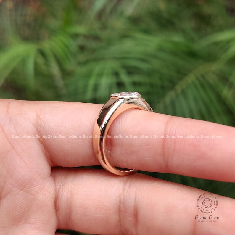 May include: A polished rose gold ring featuring a rectangular emerald-cut clear gemstone set horizontally in a bezel setting. The band has a smooth, solid metal finish with a rounded profile, showcasing a clean and simple design aesthetic.