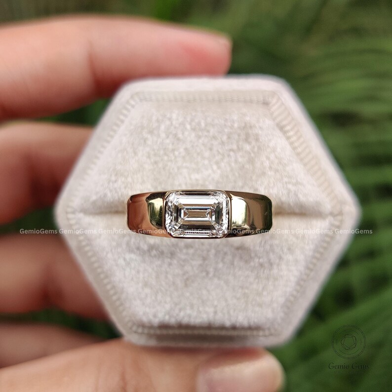 May include: A gold ring featuring a single emerald-cut clear gemstone set in a bezel setting. The band is smooth and polished, with a wide, solid design that tapers slightly toward the back. The gemstone is centered and rectangular in shape.
