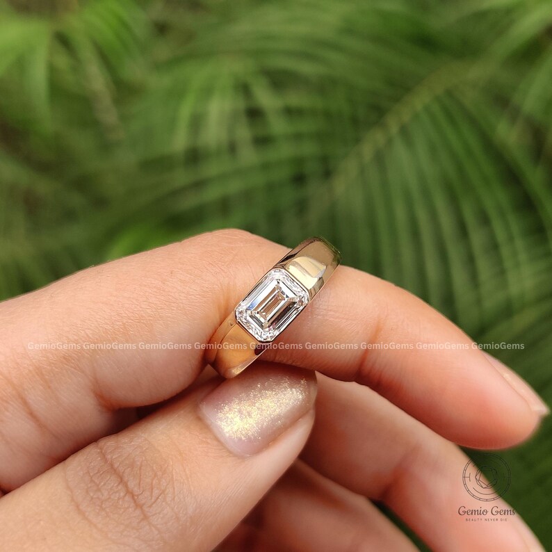 May include: A gold ring featuring a single emerald-cut diamond set in a bezel setting. The band is smooth and polished, showcasing a clean, metallic finish that complements the central rectangular gemstone. The design is simple and refined.